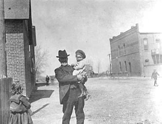 Byron North Sanford, holding grandson (Edward) Wayne Sanford. Mollie (Mary Euddra) Sanford, left. Building on right was Crawford's Saloon on ground floor, corner of Main Street and Nevada, c.1901 or 1902.